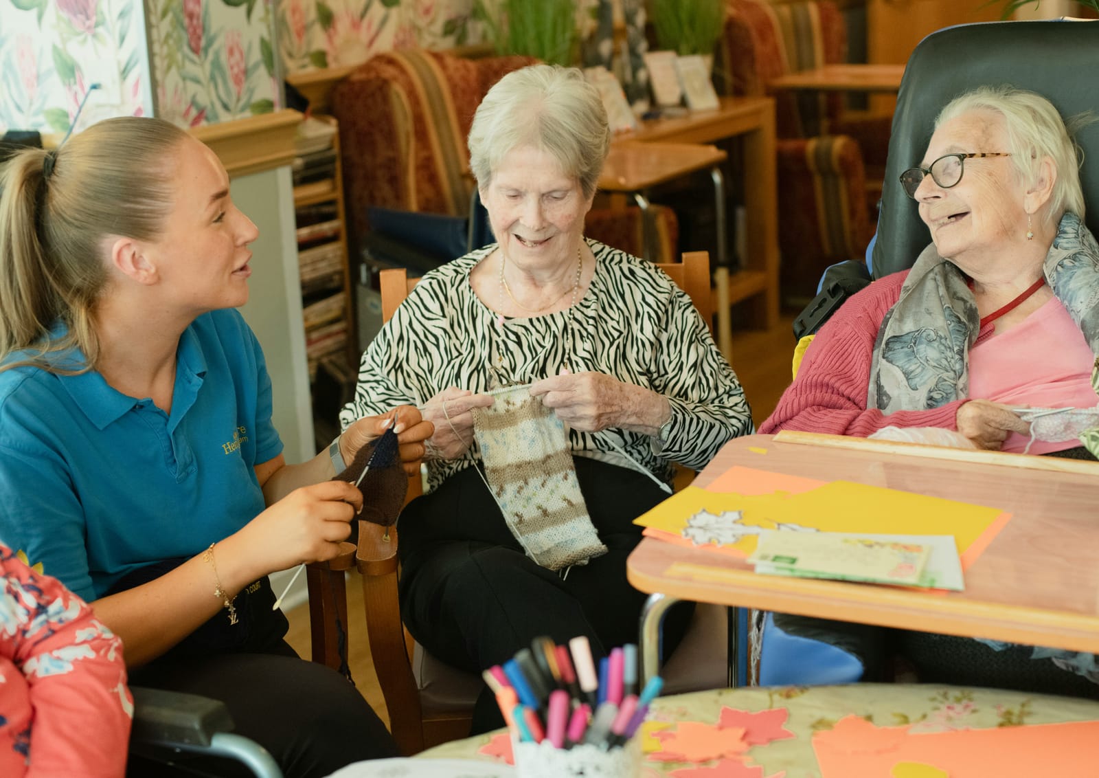 A support worker knitting alongside two residents in a care home lounge